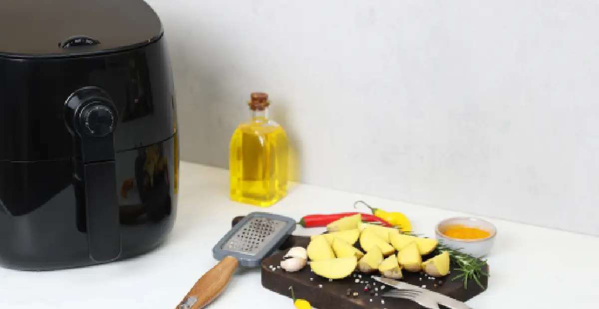 Small black air fryer beside potato wedges, oil, seasoning, and kitchen tools on a countertop